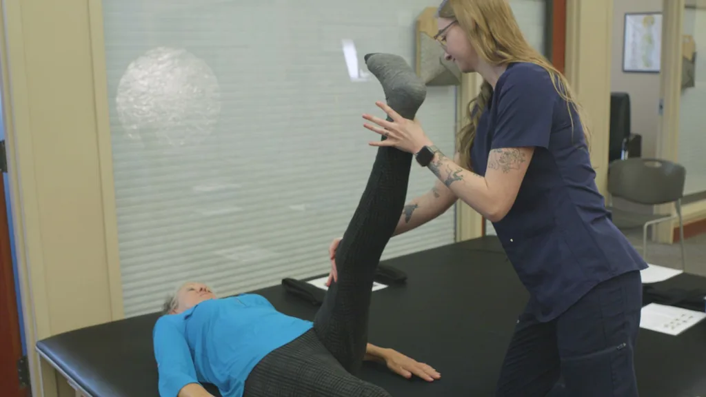A staff member guides a patient through a leg stretch on a treatment table